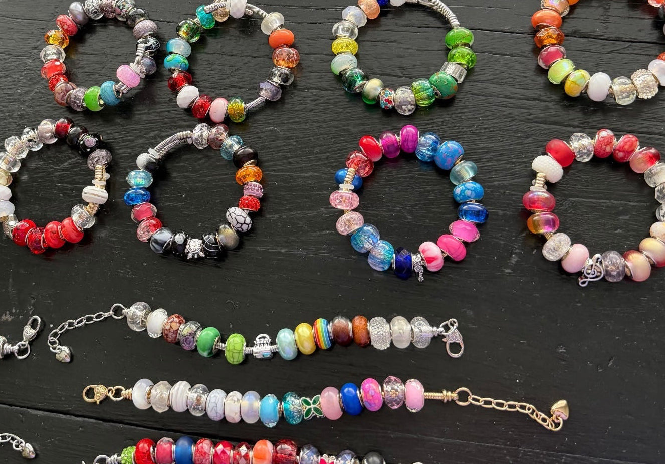A collection of colorful beaded bracelets  on a dark wooden surface. The jewelry features marbled beads in a variety of colors including pink, blue, green, yellow, orange, and white, with some pieces showing silver chain links.